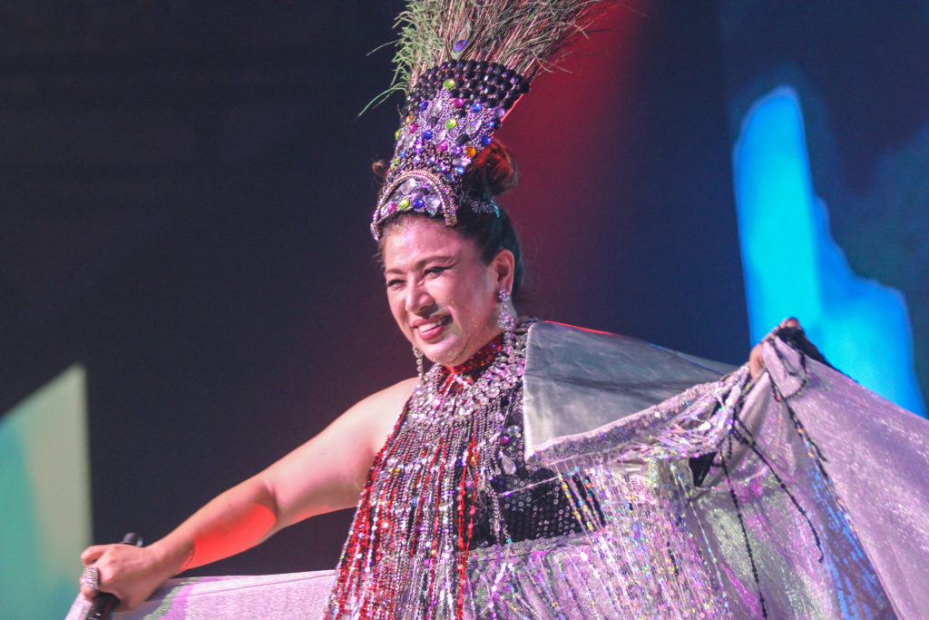 Costumed performer sings onstage under spotlight with decorative visuals.