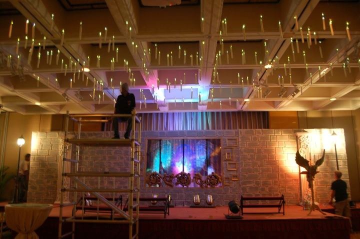 Wide view of an event hall stage setup featuring faux castle walls, floating candles being installed via scaffolding, and various props