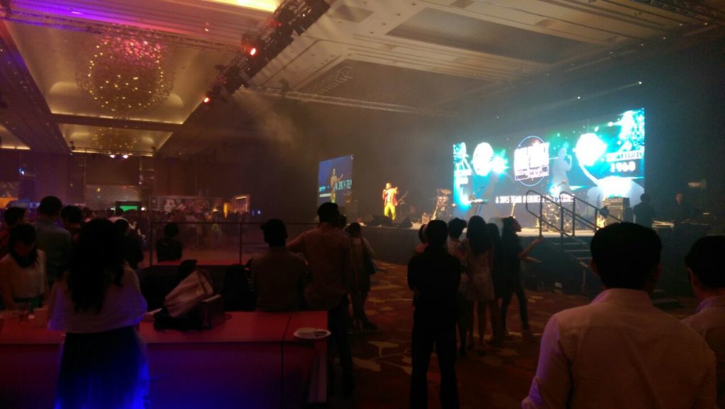 A large indoor event or concert featuring a vibrant stage setup with a performer and a massive screen displaying graphics and text.