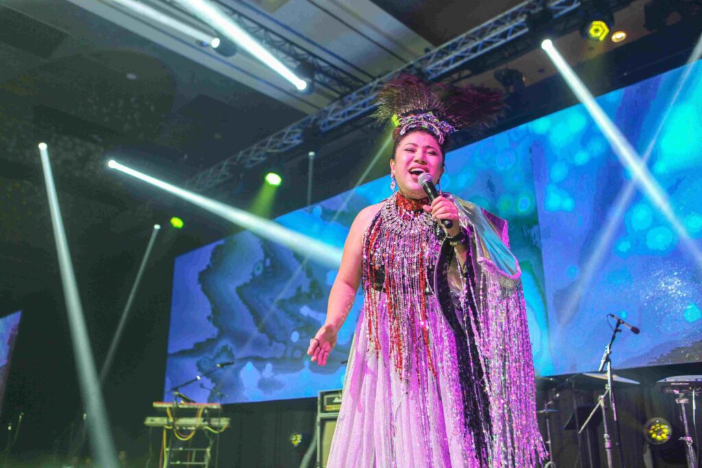Performer in a colorful costume sings on stage with abstract visuals in the background.