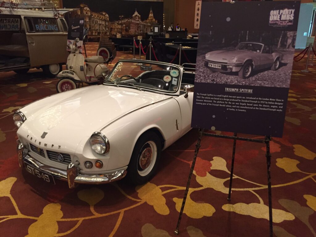 A white Triumph Spitfire displayed indoors on a patterned carpet, accompanied by a signboard detailing its history and specifications.