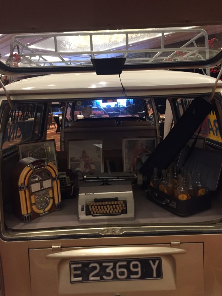 The open rear of a vintage vehicle displays a retro-themed setup featuring a jukebox on the left, a typewriter in the center, and a suitcase filled with glass bottles on the right.