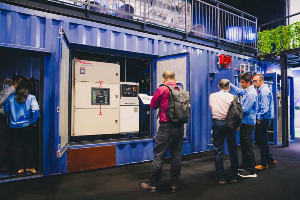 Attendees examine the open control panel of a Mitsubishi MGS2500R containerized generator at an industrial trade show.