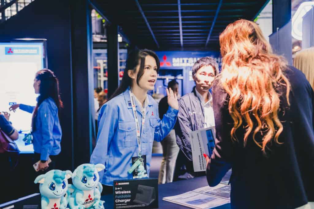 A Mitsubishi staff member welcomes visitors at the Data Centre World Asia 2025 booth, featuring SORA the Blue Dragon mascots