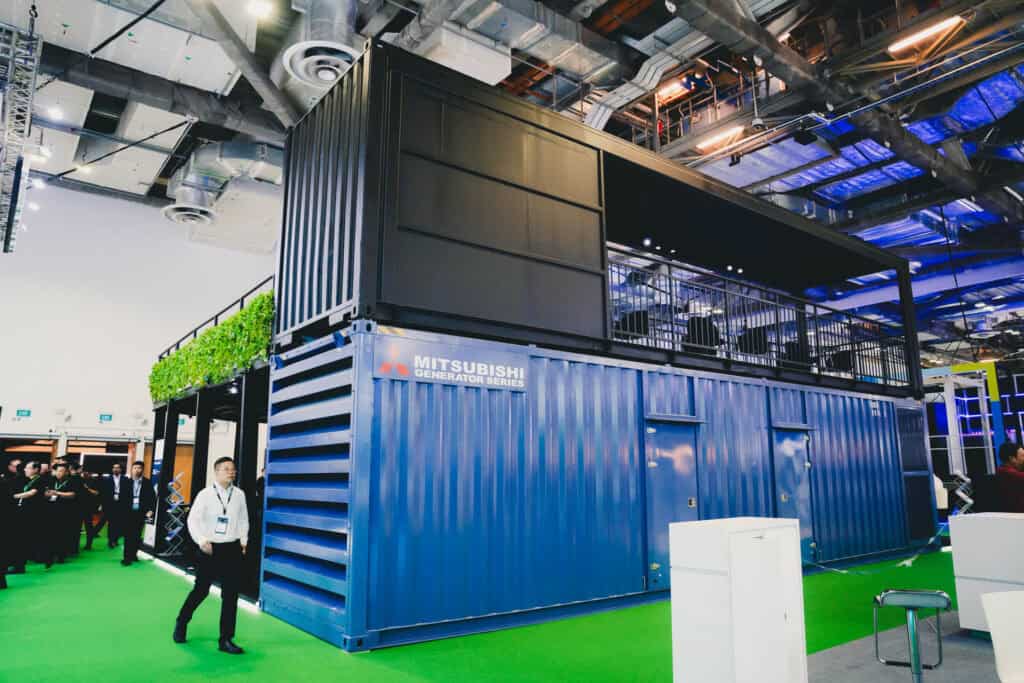 Mitsubishi Generator Series blue container unit with a black observation deck stacked on top, showcased at an industrial trade show