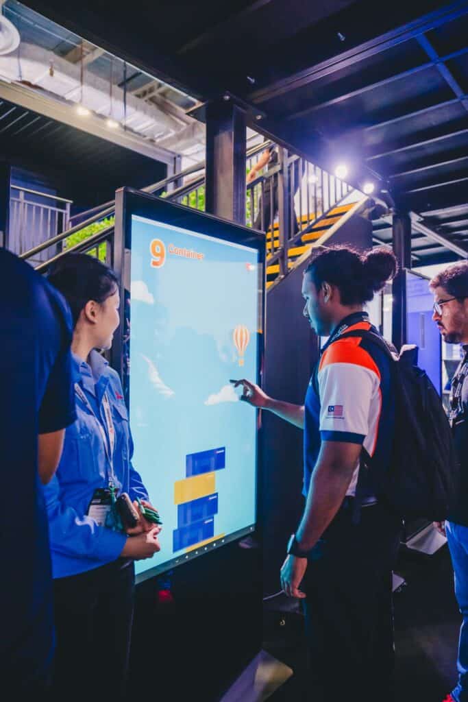 A visitor interacts with a large vertical touchscreen display featuring a Mitsubishi container-themed game at the Data Centre World Asia exhibition booth