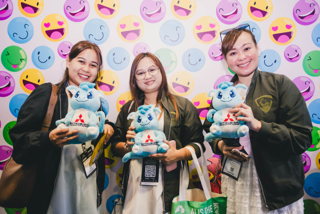 Three visitors posing with Mitsubishi 'Sora' dragon mascot plushies at the Data Centre World Asia 2025 exhibition