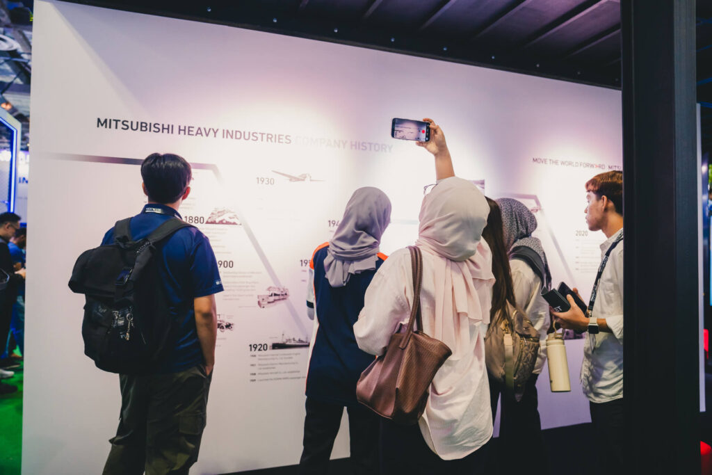 Trade show attendees explore the MHI Heritage Wall at Data Centre World Asia, featuring a visual timeline of Mitsubishi Heavy Industries' milestones and its 'Move the World Forward' mission
