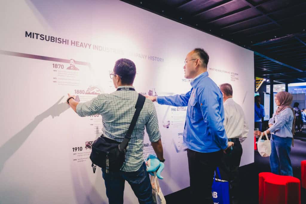 Mitsubishi staff member explaining the company's 150-year history to an attendee at a trade show.