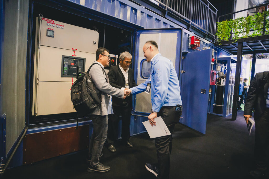 A Mitsubishi staff member shakes hands with a visitor in front of an open MGS2500R generator control panel at a trade show.