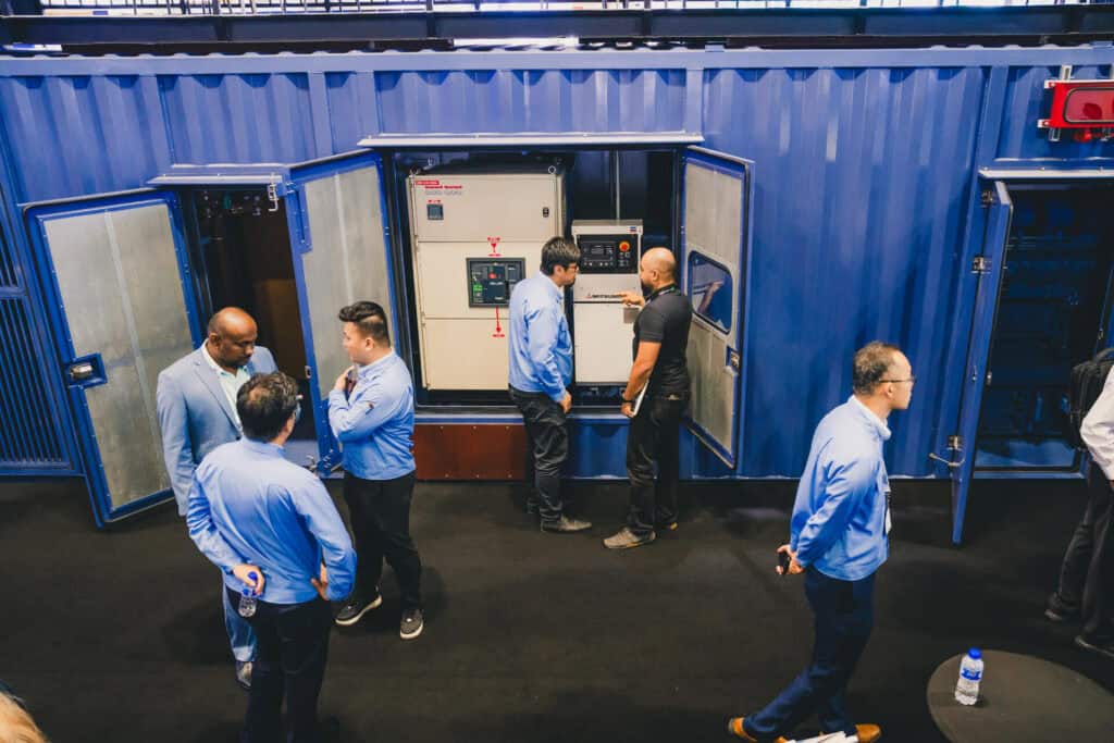 Mitsubishi representatives and trade show attendees examine the open control panel of an MGS2500R containerized generator