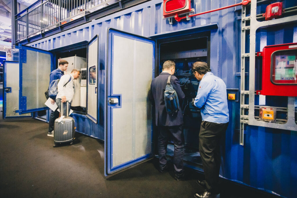 Visitors and staff inspect the internal maintenance access of a blue Mitsubishi MGS2500R containerized generator at a trade show