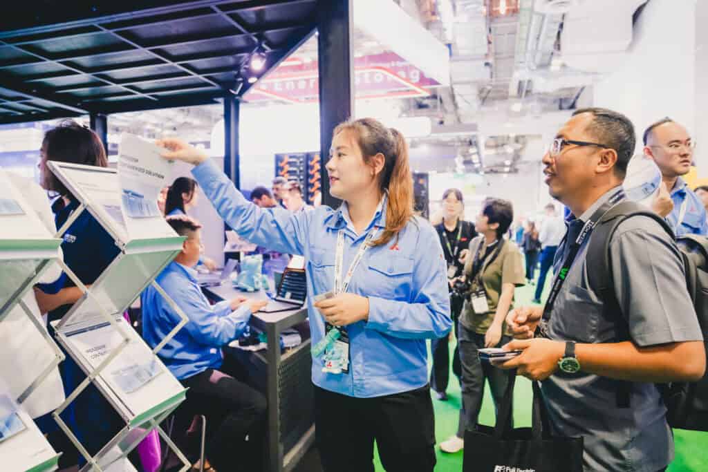 A Mitsubishi staff member handing a product brochure for the MGS2500R generator to a visitor at a trade show.