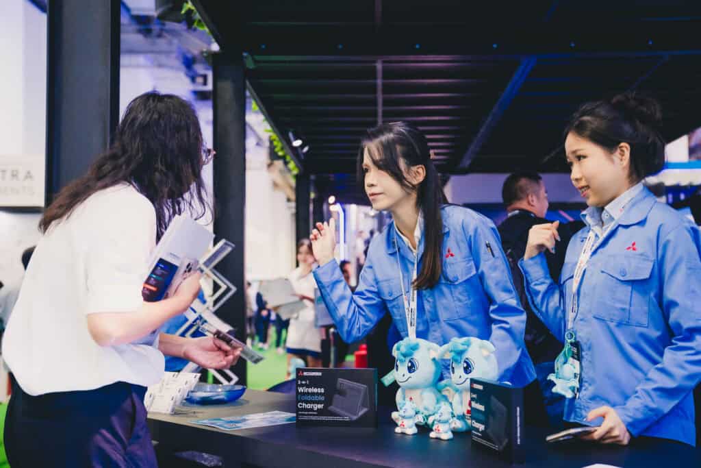 Mitsubishi staff members engaging with a visitor at their trade show booth featuring Sora mascot plushies.