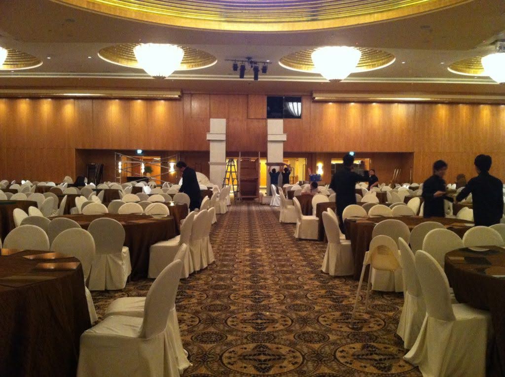 Hotel ballroom being set up for a formal event, with round tables, white chair covers, and staff arranging the venue.